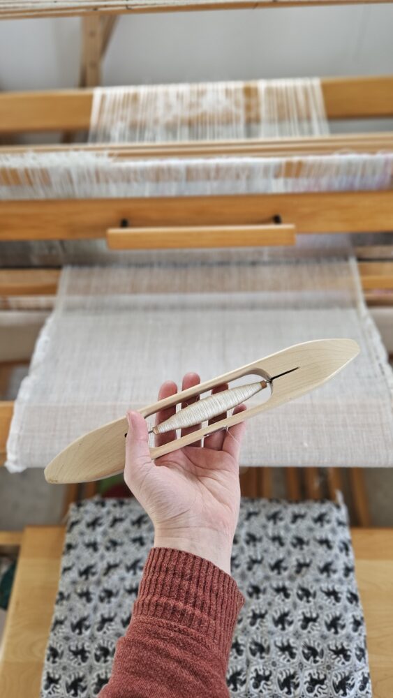 Hand holding a wooden weaving shuttle against the background of a weaving loom witgh linen kruseler fabric on it